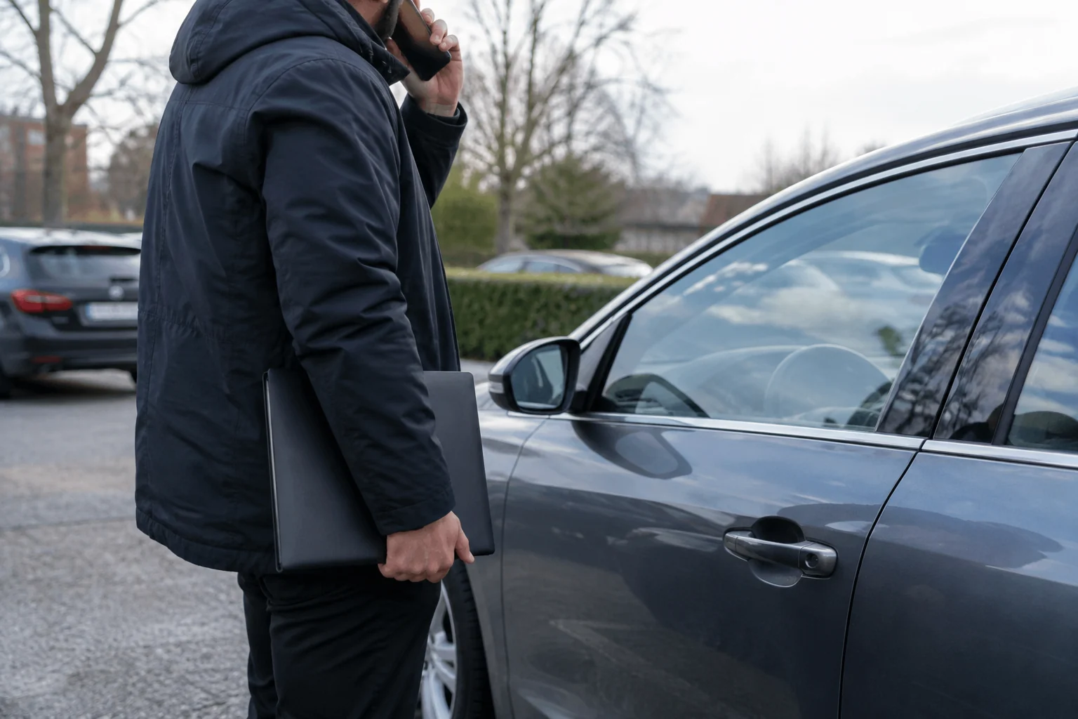 Žmogus kalba telefonu šalia automobilio, rankoje laikydamas užverstą dokumentų aplanką.